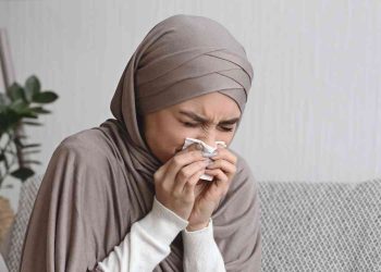 Seorang perempuan tampak bersin sambil menutup hidung dengan tisu, menggambarkan kondisi orang yang sedang kurang sehat dan membutuhkan doa serta dukungan. Dalam Islam, ungkapan syafakillah kerap disampaikan sebagai doa agar Allah memberikan kesembuhan, ketenangan, dan perlindungan bagi yang sedang sakit.