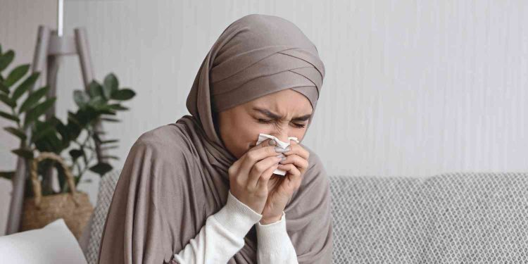Seorang perempuan tampak bersin sambil menutup hidung dengan tisu, menggambarkan kondisi orang yang sedang kurang sehat dan membutuhkan doa serta dukungan. Dalam Islam, ungkapan syafakillah kerap disampaikan sebagai doa agar Allah memberikan kesembuhan, ketenangan, dan perlindungan bagi yang sedang sakit.